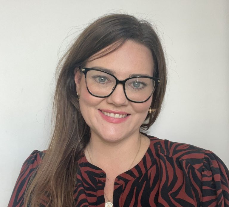 Beth McCay wearing glasses, and a patterned top against a plain background.